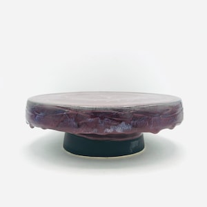 May include: A handcrafted ceramic cake stand with a dark purple, textured top and a black base. The stand is round and has a unique, artistic design. The diameter is approximately 8 inches.