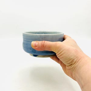 May include: A handmade ceramic bowl with a gradient of blue hues. The bowl is held in a hand, showcasing its rounded shape and textured surface. The bowl's base is a lighter shade, transitioning to a deeper blue towards the rim.