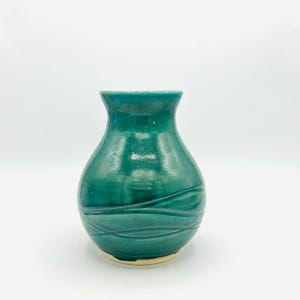 May include: A small, teal-colored ceramic vase with a rounded body and a narrow neck. The vase features a series of horizontal, wave-like grooves around its midsection. The base is a light beige color. The vase is set against a white background.