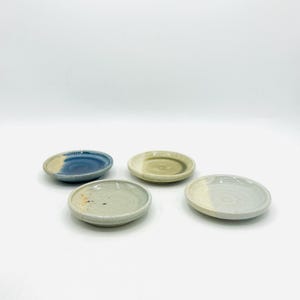 May include: Four small, handmade ceramic dishes in shades of blue, green, and beige. Each dish has a unique color pattern and a slightly irregular shape, suggesting a handcrafted origin. The dishes are arranged on a white surface.