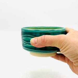 May include: A teal ceramic bowl with a textured wave pattern on the side. The bowl has a cream-colored base and is held by a hand against a white background. The bowl is likely used for serving food or beverages.