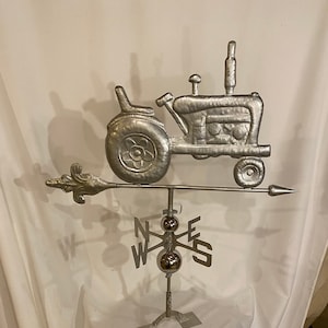 May include: A silver metal tractor weather vane with a compass rose and a fleur-de-lis finial. The tractor is facing right and has a detailed design.