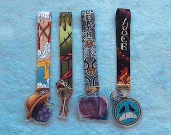 May include: Four fabric wristlet keychains featuring One Piece anime designs. The first has a straw hat and fruit charm, the second a sword charm, the third a heart-shaped charm with the word DEATH, and the fourth a circular blue face charm with ACE.