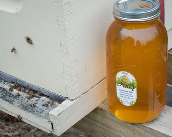 Puede incluir: Un gran tarro de vidrio lleno de miel dorada, etiquetado como "Pure Honey". El tarro está junto a una colmena de madera blanca, con varias abejas visibles. La etiqueta incluye el texto "How Sweet It Is".