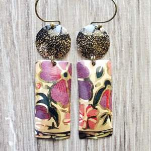May include: A pair of dangle earrings featuring a circular top piece with a black and gold speckled finish, connected to a rectangular bottom piece. The rectangular section displays a floral pattern in shades of purple, pink, and black on a gold background.