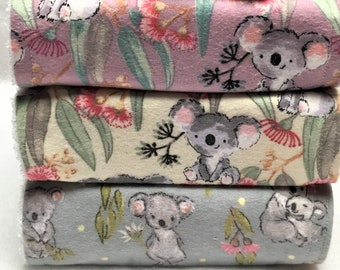 May include: A grey koala soft toy sits on a stack of three fabric-covered blocks. The blocks feature koala and floral patterns in pink, beige, and grey. The koala has large, round eyes and a black nose.