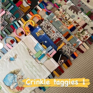 May include: A collection of colorful crinkle taggies with various patterns, including foxes, bears, and floral designs. Each taggie features a ribbon and a plastic ring. The image also includes a card that says "A gift for you..." and the text "Crinkle taggies 1".