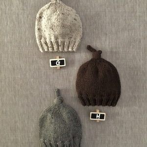 May include: Three knitted baby hats in different colours: beige, brown, and grey. Each hat has a knot on top and is labelled with a letter: G, H, and I.