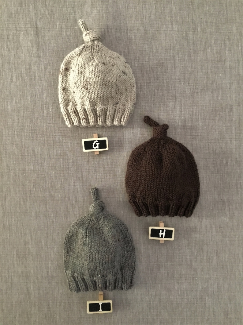 May include: Three knitted baby hats in different colours: beige, brown, and grey. Each hat has a knot on top and is labelled with a letter: G, H, and I.