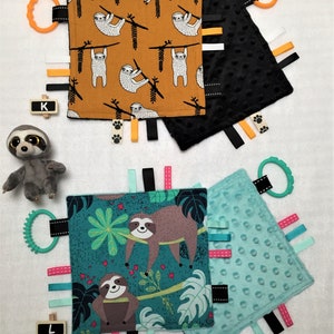 May include: A collection of baby sensory tag blankets with sloth designs. One blanket features sloths on a mustard background, another has a black textured surface, and the third has a teal background with sloths and tropical leaves. Each has colorful ribbons and a teether.