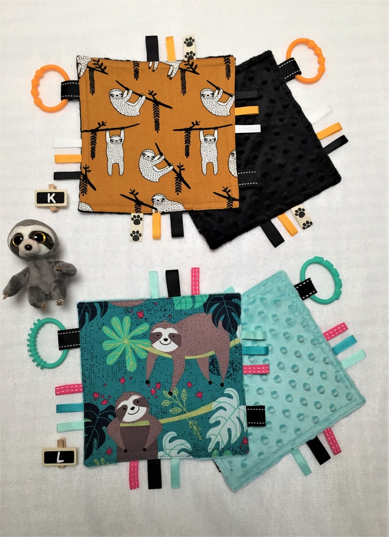 May include: A collection of baby sensory tag blankets with sloth designs. One blanket features sloths on a mustard background, another has a black textured surface, and the third has a teal background with sloths and tropical leaves. Each has colorful ribbons and a teether.
