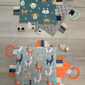 May include: Two baby sensory blankets. One teal blanket features dog faces and paw prints, with a green teether ring. The other blanket has a blue background with llama designs and an orange teether ring. Both have ribbons.