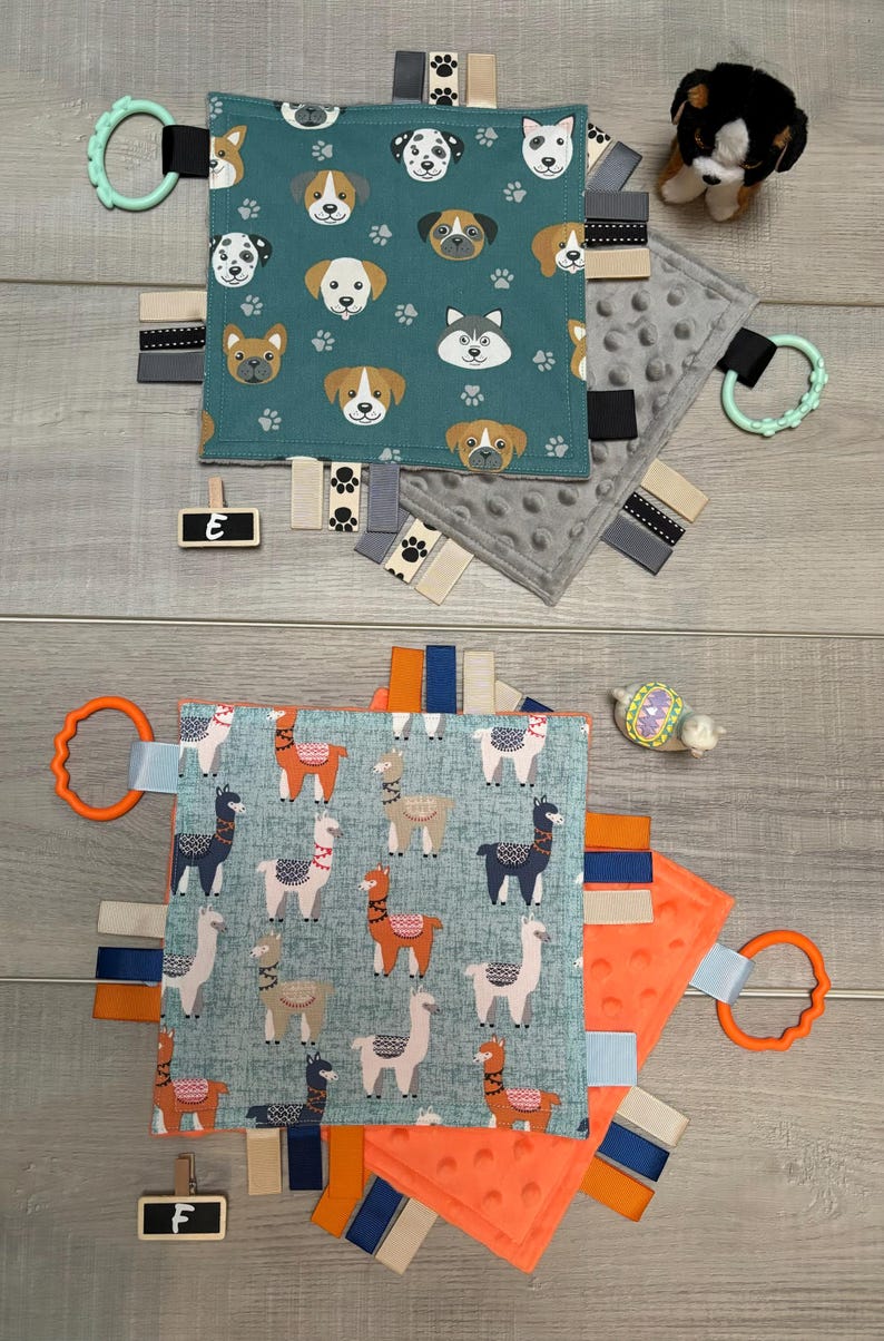 May include: Two baby sensory blankets. One teal blanket features dog faces and paw prints, with a green teether ring. The other blanket has a blue background with llama designs and an orange teether ring. Both have ribbons.