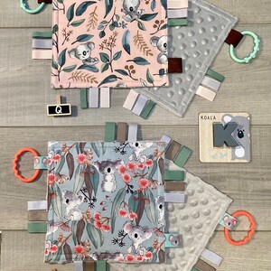 May include: Baby sensory squares with koala bear designs. One square features a pink background with koalas and foliage, the other a blue background with koalas and flowers. Each has ribbons and a teether.