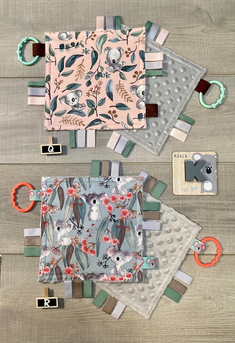 May include: Baby sensory squares with koala bear designs. One square features a pink background with koalas and foliage, the other a blue background with koalas and flowers. Each has ribbons and a teether.