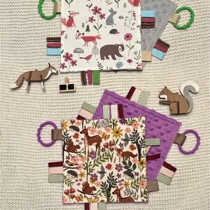 May include: Two baby sensory tag blankets with woodland animal patterns, featuring foxes, bears, and deer. Each blanket has colorful ribbons, a textured backing, and a plastic ring. Wooden animal cutouts and a small sign with the letter "J" are also present.