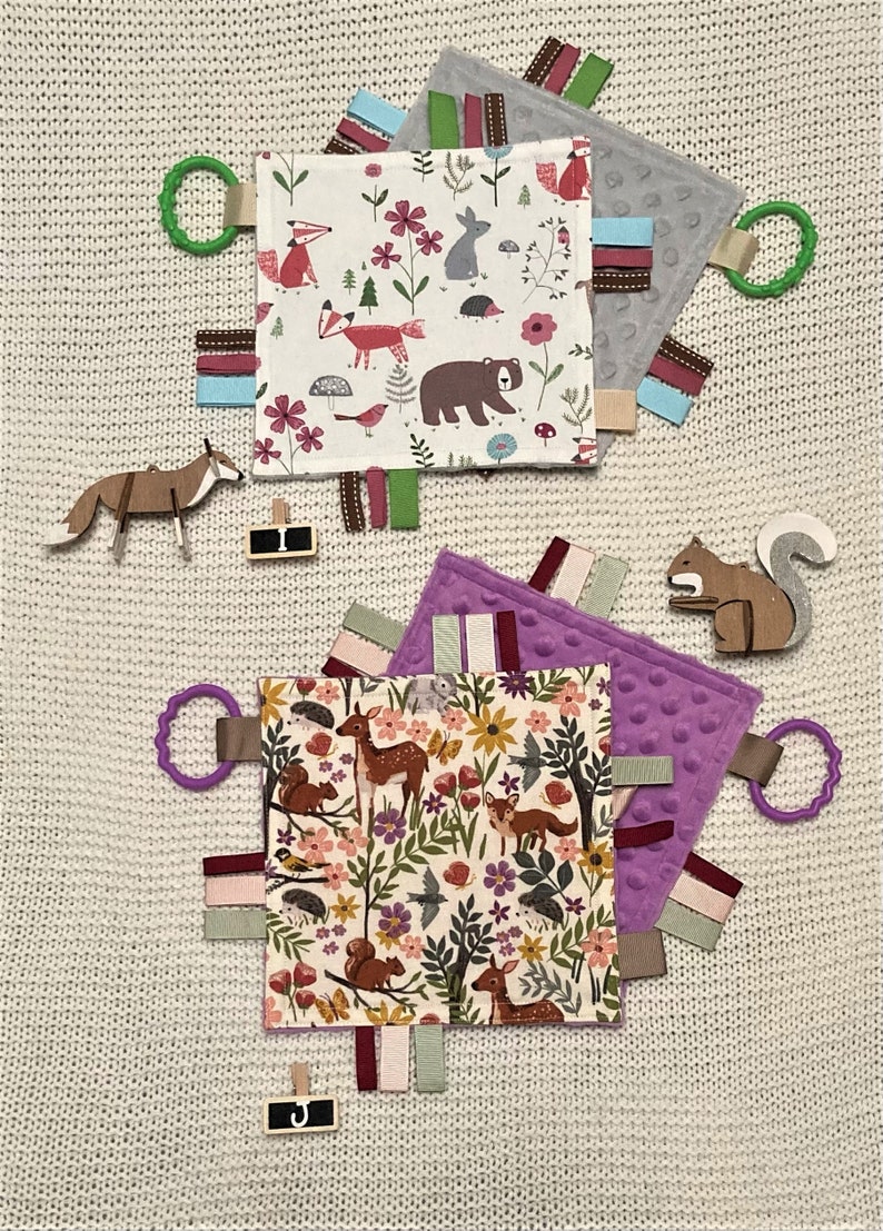 May include: Two baby sensory tag blankets with woodland animal patterns, featuring foxes, bears, and deer. Each blanket has colorful ribbons, a textured backing, and a plastic ring. Wooden animal cutouts and a small sign with the letter "J" are also present.