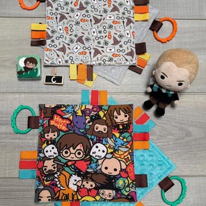 May include: Two baby sensory blankets with Harry Potter-themed designs. One features a repeating pattern of icons, the other has a colorful cartoon graphic. Each blanket has a textured backing, colorful ribbons, and a plastic ring. A small plush figure and block are also present.