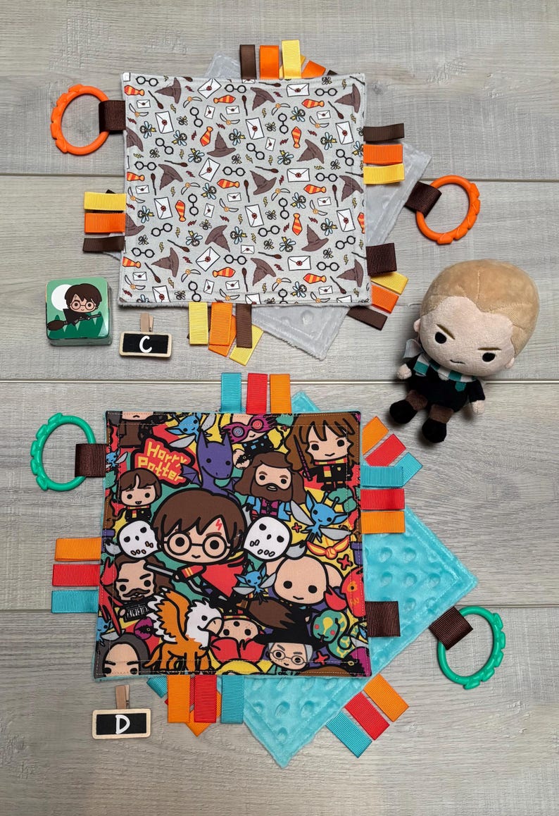 May include: Two baby sensory blankets with Harry Potter-themed designs. One features a repeating pattern of icons, the other has a colorful cartoon graphic. Each blanket has a textured backing, colorful ribbons, and a plastic ring. A small plush figure and block are also present.