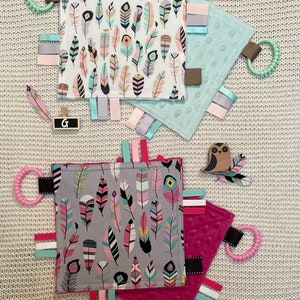 May include: Three baby tag blankets with feather patterns in pink, teal, and gray. Each blanket has a textured surface, colorful ribbons, and a teething ring. Decorative wooden feather and owl accents are also present.