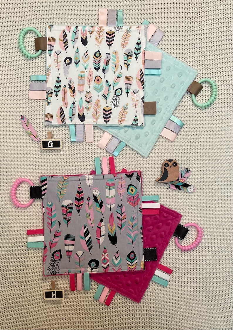 May include: Three baby tag blankets with feather patterns in pink, teal, and gray. Each blanket has a textured surface, colorful ribbons, and a teething ring. Decorative wooden feather and owl accents are also present.