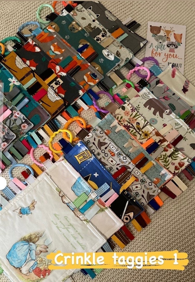 May include: A collection of colorful crinkle taggies with various patterns, including foxes, bears, and floral designs. Each taggie features a ribbon and a plastic ring. The image also includes a card that says "A gift for you..." and the text "Crinkle taggies 1".