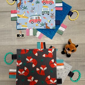 May include: Two baby sensory blankets with attached ribbons and teething rings. One blanket has a blue background with koalas, surfboards, and vans. The other has a gray background with red foxes. A wooden koala puzzle is also present.
