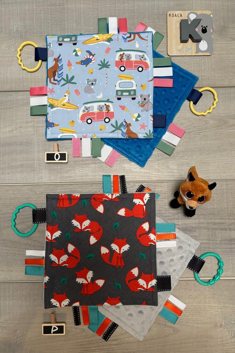 May include: Two baby sensory blankets with attached ribbons and teething rings. One blanket has a blue background with koalas, surfboards, and vans. The other has a gray background with red foxes. A wooden koala puzzle is also present.