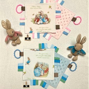 May include: Two baby security blankets with attached ribbons and a plush bunny rabbit. The blankets feature illustrations of Peter Rabbit and text from the story. One blanket is pink, the other is blue. A pink and a blue ring are attached to the blankets.