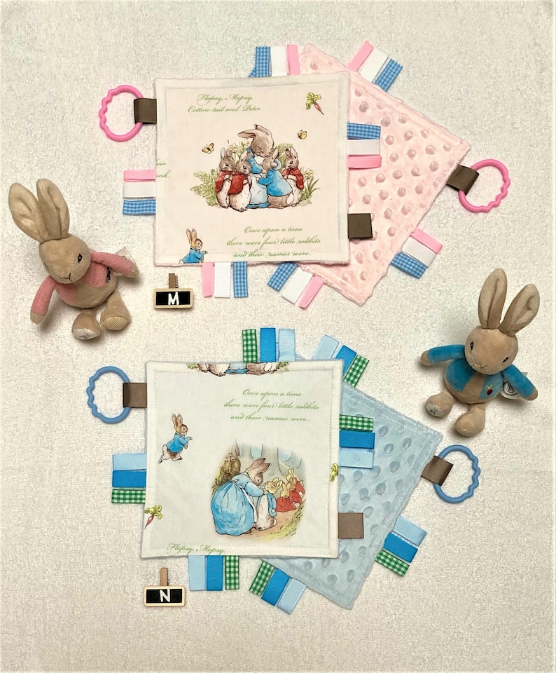 May include: Two baby security blankets with attached ribbons and a plush bunny rabbit. The blankets feature illustrations of Peter Rabbit and text from the story. One blanket is pink, the other is blue. A pink and a blue ring are attached to the blankets.