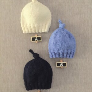 May include: Three knitted baby hats in different colours: white, light blue, and dark blue. Each hat has a knot on top and is labelled with a letter A, B, and C.