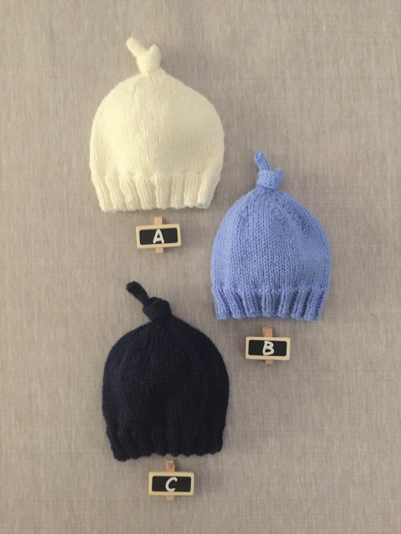 May include: Three knitted baby hats in different colours: white, light blue, and dark blue. Each hat has a knot on top and is labelled with a letter A, B, and C.