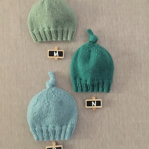 May include: Three knitted hats in different shades of green. The hats have a knot on top and are labelled with the letters M, N, and O.