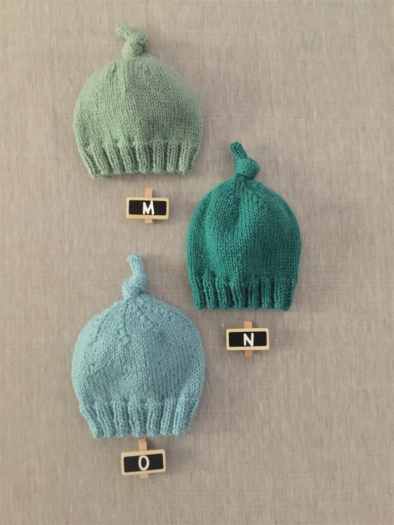 May include: Three knitted hats in different shades of green. The hats have a knot on top and are labelled with the letters M, N, and O.