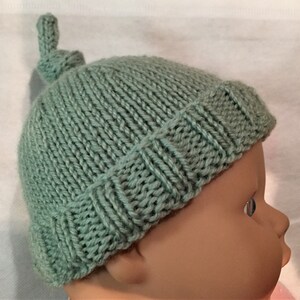 May include: A soft, sage green knitted baby hat with a knotted top. The hat is made with a simple stitch pattern and has a fold-over brim.