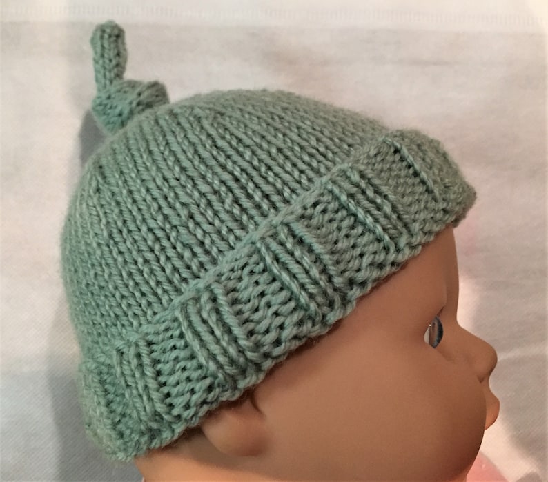 May include: A soft, sage green knitted baby hat with a knotted top. The hat is made with a simple stitch pattern and has a fold-over brim.
