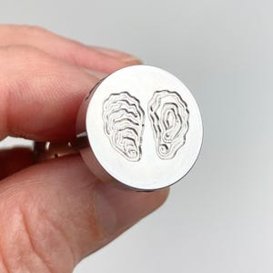 May include: A circular silver-toned metal cufflink featuring an engraved design of two oyster shells side by side on the flat face. The surface of the metal is smooth and reflective, with the oyster shell details etched in a darker, recessed tone.