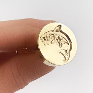 May include: A gold-toned metal seal with a detailed design of a stylized shark. The seal is round and has a smooth surface.