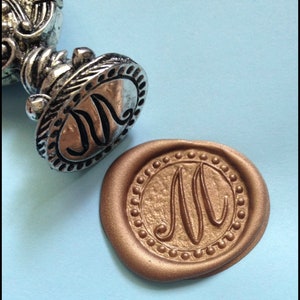 May include: A silver wax seal stamp with the letter "M" engraved on it. The stamp is used to create a wax seal with the same letter, which is shown in the image. The wax seal is a circular shape with a gold color.
