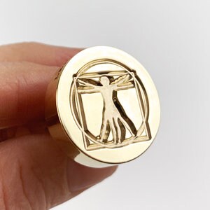 May include: Gold-toned metal ring with a raised design of a human figure inside a square and circle. The design is reminiscent of Leonardo da Vinci's Vitruvian Man.