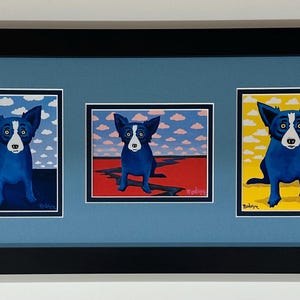 May include: Framed artwork featuring three paintings of a blue dog with a white face and yellow eyes. Each painting has a different background: blue sky with clouds, red ground, and yellow background. The artwork is framed in black.