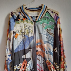 Könnte beinhalten: Eine schwarze Bomberjacke mit Reißverschluss und einem bunten Patchwork-Design mit japanisch inspirierten Mustern. Die Jacke hat einen gerippten Kragen, Bündchen und Saum mit blauen und weißen Streifen.