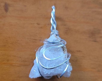 May include: A clear quartz crystal wrapped in silver wire, forming a pendant. The crystal is irregularly shaped and has a rough texture. The wire is twisted and forms a delicate cage around the crystal.