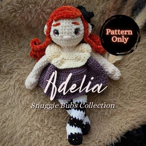 May include: A crocheted doll named Adelia, with red hair, a purple dress, and black and white striped socks. The doll has a cream-colored collar and is part of the Snuggle Bubs' Collection. The image includes a circular graphic that says "Pattern Only".
