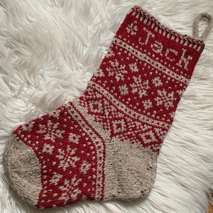 Peut inclure: Une chaussette de Noël tricotée rouge et beige avec un motif de flocon de neige et le nom "Jack" tricoté en haut. La chaussette a une boucle pour l'accrocher et repose sur une surface blanche et moelleuse.