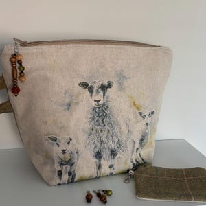 May include: A beige fabric zipped pouch featuring a printed illustration of a sheep and two lambs. The pouch includes a decorative beaded pull charm. Beside it sits a smaller, rectangular brown and blue plaid fabric pouch and two loose beads.