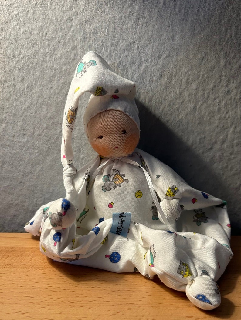 Pu&ograve; includere: Bambola di stoffa bianca con un cappello di stoffa bianca con un motivo colorato di animali e stelle. La bambola indossa un sacco a pelo di stoffa bianca con un motivo colorato di animali e stelle. La bambola ha un'etichetta di stoffa bianca con il testo "d'Amiens" su di essa.