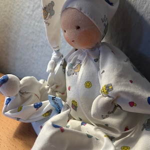 Pu&ograve; includere: Una bambola di stoffa bianca con un cappello di stoffa bianca. La bambola indossa una tutina bianca con un motivo colorato di animali e cuori. La bambola &egrave; seduta su una superficie di legno.