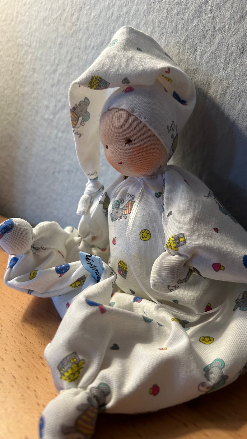 Pu&ograve; includere: Una bambola di stoffa bianca con un cappello di stoffa bianca. La bambola indossa una tutina bianca con un motivo colorato di animali e cuori. La bambola &egrave; seduta su una superficie di legno.