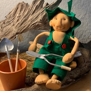 Pode incluir: Um gnomo de pelúcia vestindo um chapéu verde e macacão, segurando uma tesoura. O gnomo está sentado em um pedaço de madeira flutuante com um pequeno pote contendo ferramentas de jardinagem.